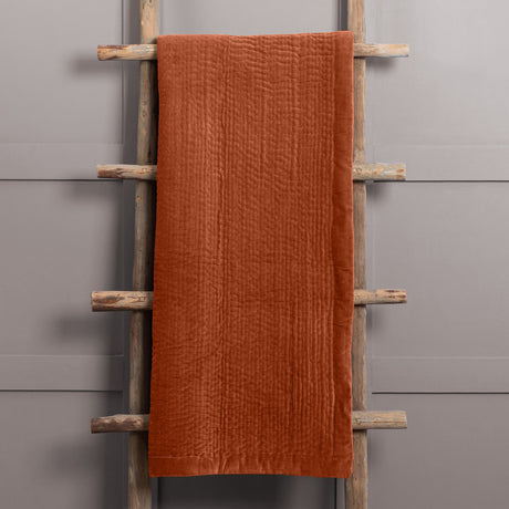 Haze Velvet Cotton Throw in Sunset, a rust-orange quilted cotton velvet throw with rippled channel texture, draped over a wooden ladder against a grey background.