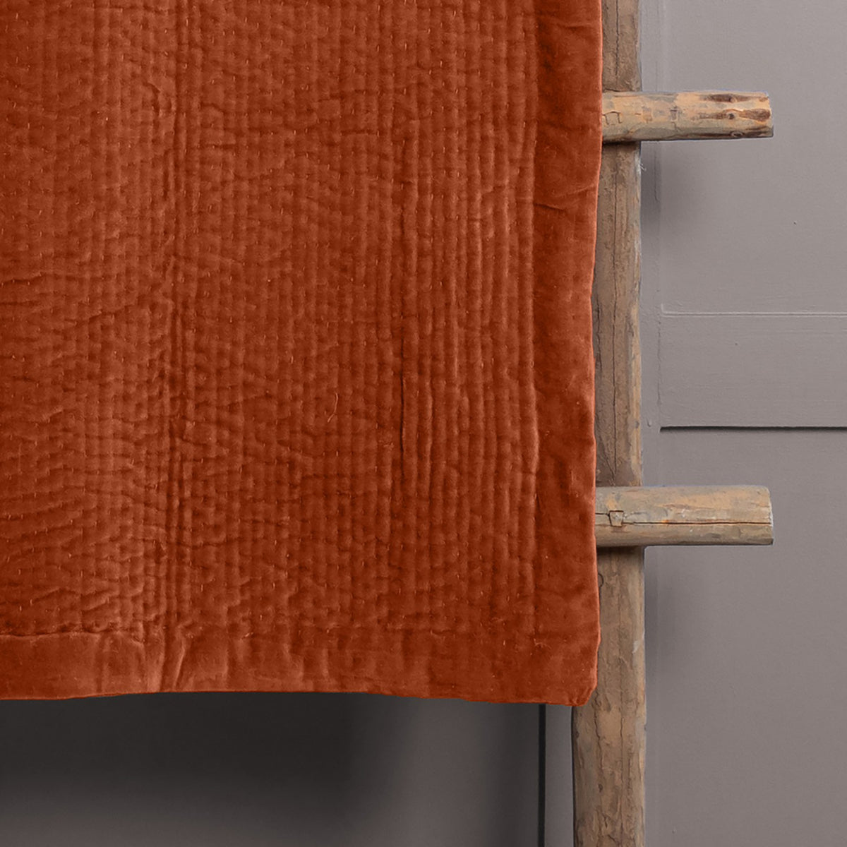 Haze Velvet Cotton Throw in rust-orange, hand-quilted cotton velvet with rippled channel texture, draped over a rustic wooden ladder against a grey wall.