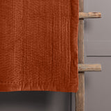 Haze Velvet Cotton Throw in rust-orange, hand-quilted cotton velvet with rippled channel texture, draped over a rustic wooden ladder against a grey wall.