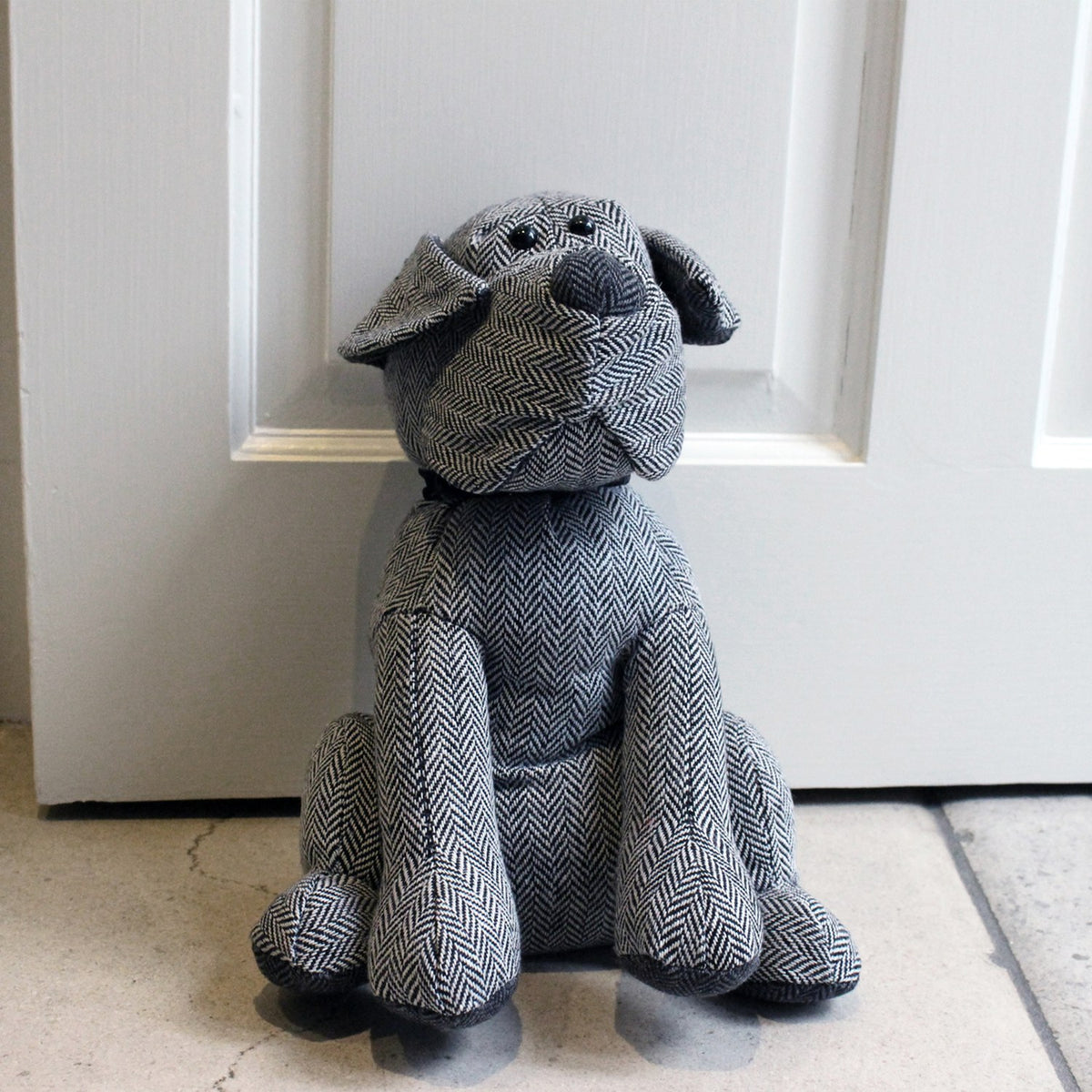 Herringbone Dog Animal Polyester Door Stop in Grey, sitting dog shape with black collar, beaded eyes and darker ears, paws and nose, set against a white door.