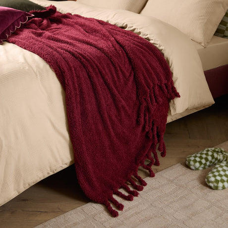 Harlan Borg Sherpa Plain Polyester Throw in burgundy, draped over a bed with tasselled fringe edge, styled with cream bedding in a bedroom setting.