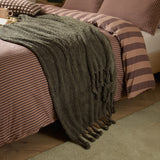 Harlan Borg Sherpa Plain Polyester Throw in olive green draped over a striped bed, showing its curly-piled texture and tasselled fringe edge.