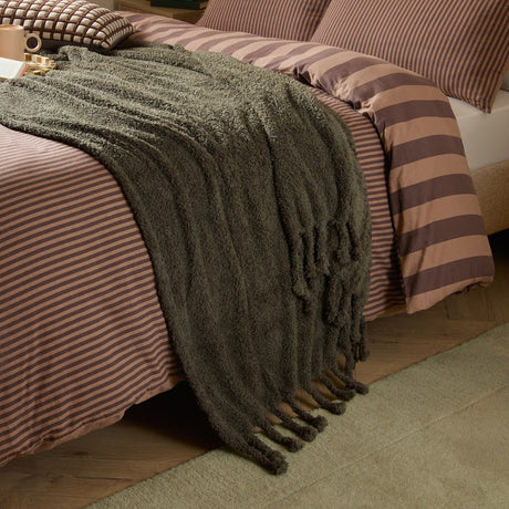 Harlan Borg Sherpa Plain Polyester Throw in olive green draped over a striped bed, showing its curly-piled texture and tasselled fringe edge.