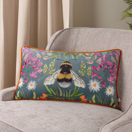 Rectangular velvet cushion from the House of Bloom range with a hand-painted bumble bee and pink, white and orange florals on a blue-grey ground, shown on a beige armchair.