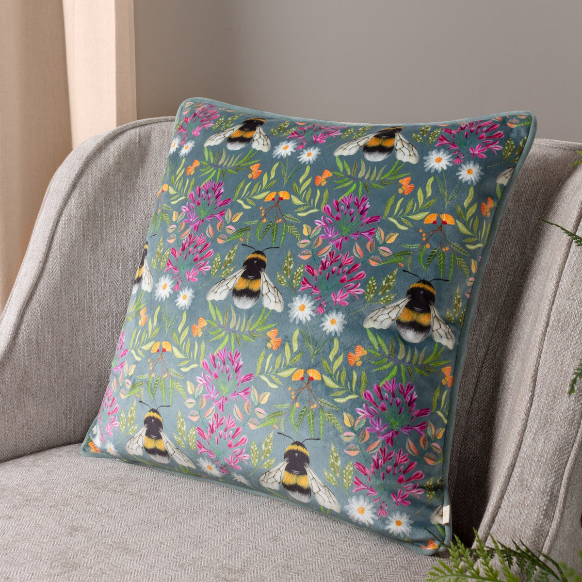 House of Bloom Floral Velvet Cushion in blue-grey, printed with bumble bees and pink florals, shown on a grey armchair.
