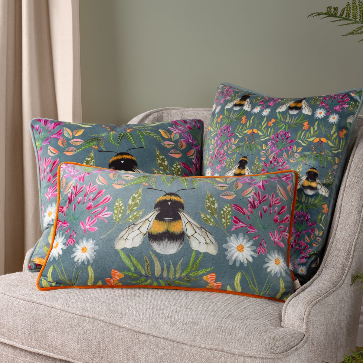 House of Bloom Floral Velvet Cushions in blue-grey with bumble bee and pink floral print, one rectangular with orange piping, styled on a beige armchair.