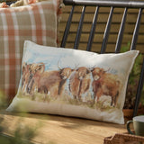 Rectangular linen-cotton cushion featuring a hand-painted watercolour highland cattle design in russet and amber, shown against a checked cushion on an outdoor bench.