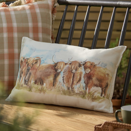 Rectangular linen-cotton cushion featuring a hand-painted watercolour highland cattle design in russet and amber, shown against a checked cushion on an outdoor bench.