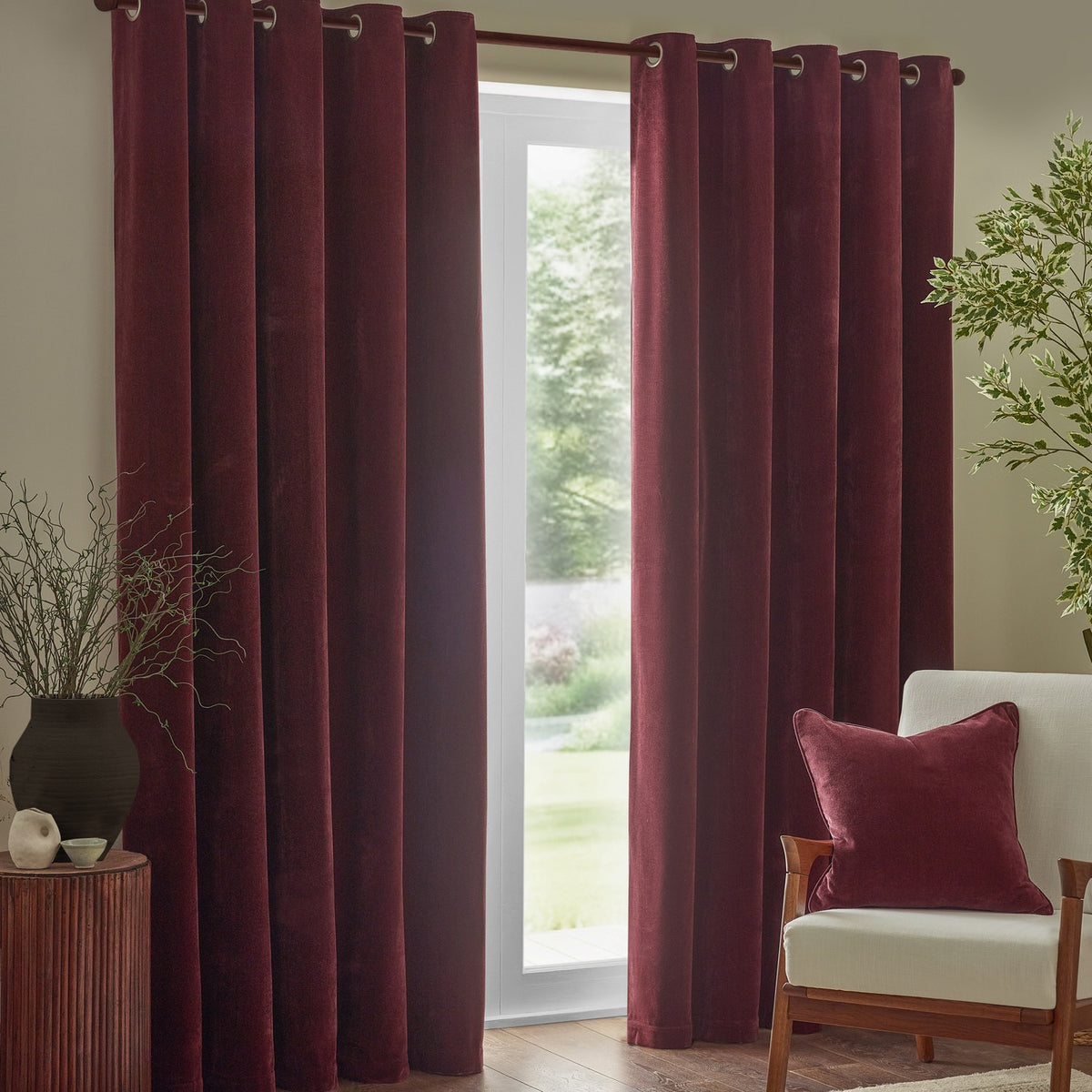 Heavy chenille velvet eyelet curtains in beetroot red hanging in full folds beside a glass door, with a matching cushion on a cream armchair in a living room.