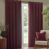 Heavy chenille velvet eyelet curtains in beetroot red hanging in full folds beside a glass door, with a matching cushion on a cream armchair in a living room.