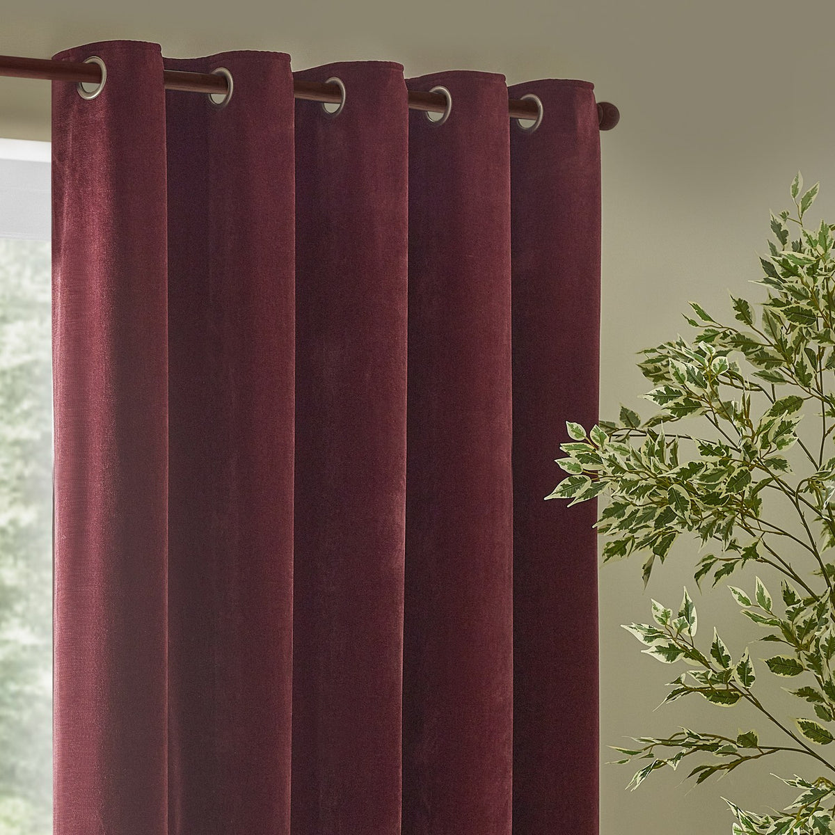 Heavy chenille velvet eyelet curtains in deep beetroot red, hanging in rounded folds with silver-toned eyelet rings on a curtain pole, near a window and green foliage.