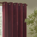 Heavy chenille velvet eyelet curtains in deep beetroot red, hanging in rounded folds with silver-toned eyelet rings on a curtain pole, near a window and green foliage.