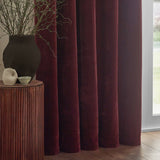 Heavy chenille velvet eyelet curtains in deep beetroot red, shown in full rounded folds by a window, styled with a dark vase and wooden side table.