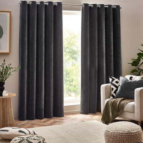 Charcoal chenille velvet eyelet curtains hanging in deep folds either side of a living room window, shown with cream armchair, cushions, and wooden furniture.