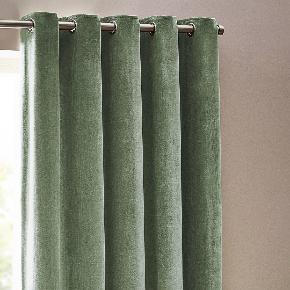 Heavy chenille velvet eyelet curtains in eucalyptus green, shown hanging in rounded folds from a metal pole in a room with light walls.