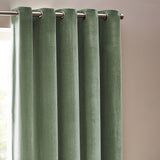 Heavy chenille velvet eyelet curtains in eucalyptus green, shown hanging in rounded folds from a metal pole in a room with light walls.