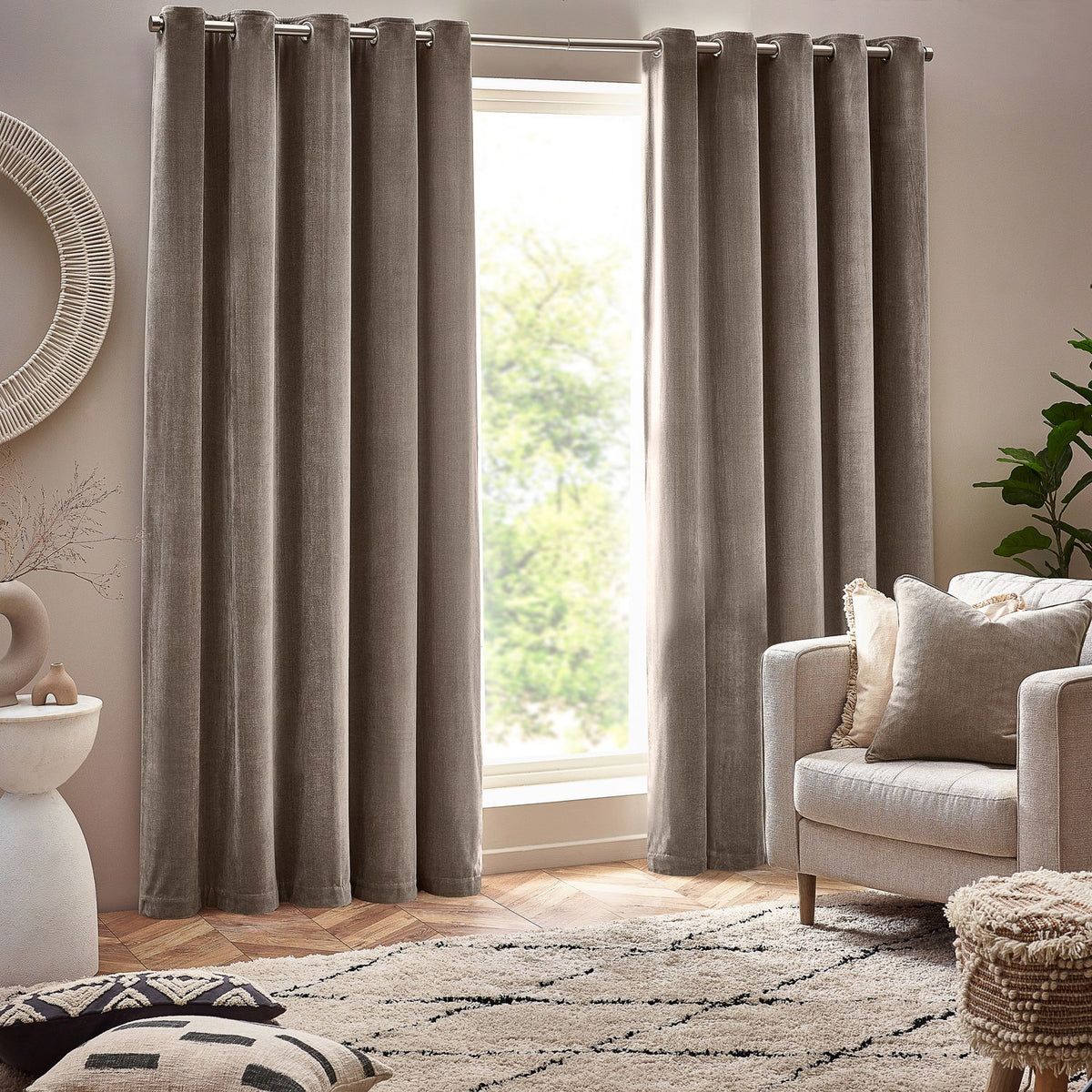 Heavy chenille velvet lined eyelet curtains in greige, hung floor-length either side of a window in a living room with an armchair and patterned rug.