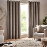 Heavy chenille velvet lined eyelet curtains in greige, hung floor-length either side of a window in a living room with an armchair and patterned rug.