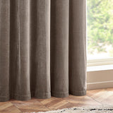 Heavy chenille velvet lined eyelet curtains in greige, shown in soft folds beside a window with wood flooring in a living room setting.