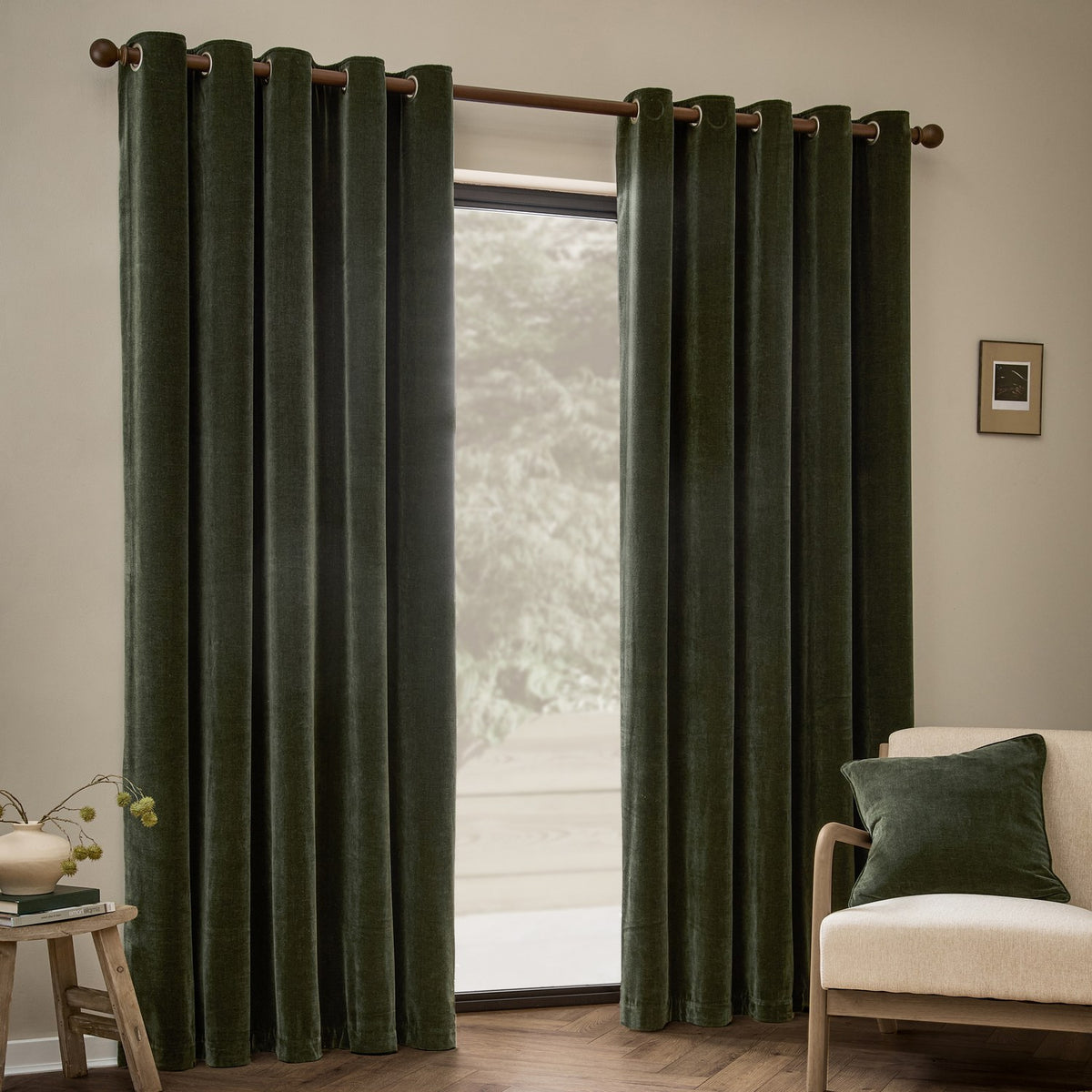 Heavy Chenille Velvet Fully Lined Eyelet Curtains in lichen green hung on a wooden pole beside a window, shown in a living room with a cream armchair and matching cushion.