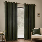 Heavy Chenille Velvet Fully Lined Eyelet Curtains in lichen green hung on a wooden pole beside a window, shown in a living room with a cream armchair and matching cushion.