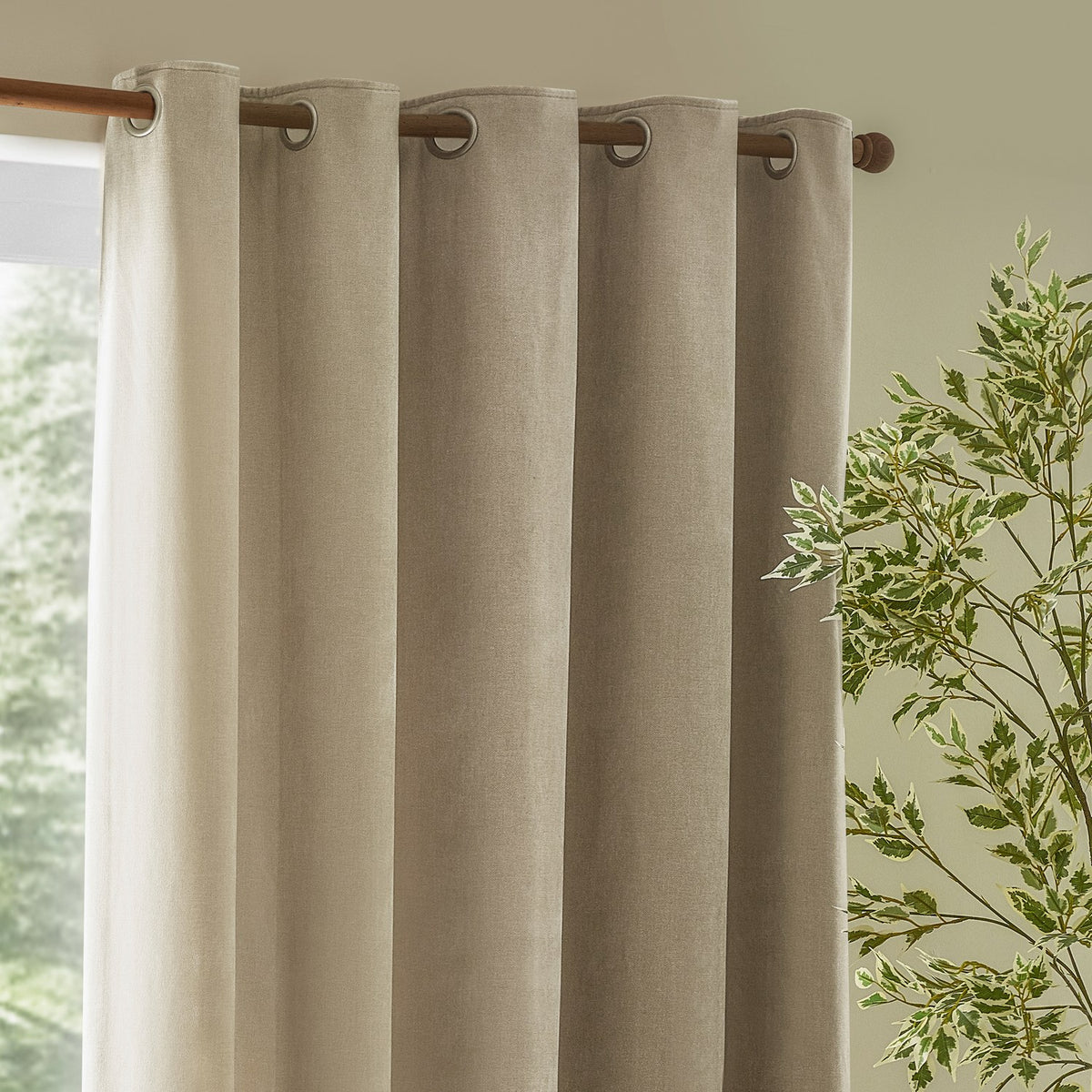 Heavy Chenille Linen Lined Eyelet Curtains in linen, hung on a wooden pole with metal eyelets in a living room beside a window and green foliage.