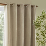 Heavy Chenille Linen Lined Eyelet Curtains in linen, hung on a wooden pole with metal eyelets in a living room beside a window and green foliage.