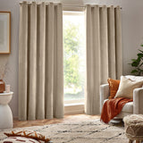 Natural beige heavy chenille velvet eyelet curtains hung either side of a window in a living room, styled with an armchair, cushions and patterned rug.
