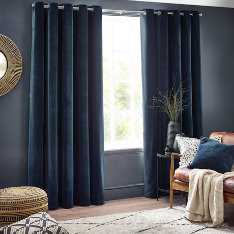 Navy chenille velvet eyelet curtains hanging on a metal rod by a window, styled in a living room with a leather armchair, side table, and patterned rug.