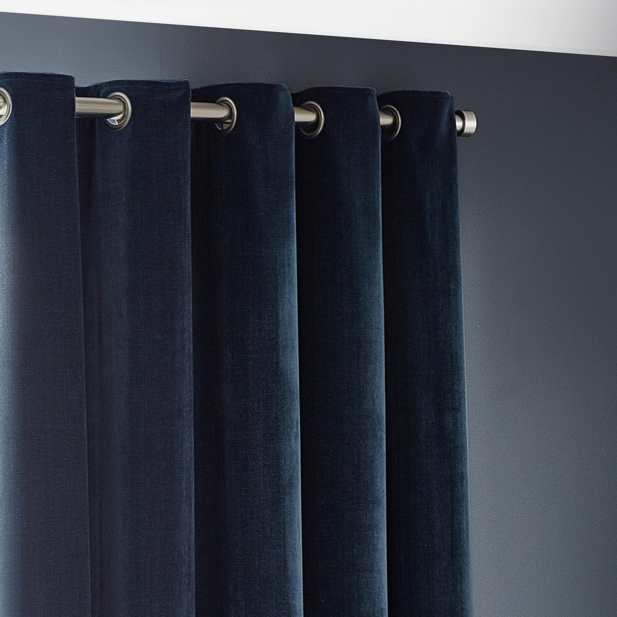 Close-up of navy chenille velvet eyelet curtains with brushed metal grommet rings hung on a matching pole against a dark grey wall.