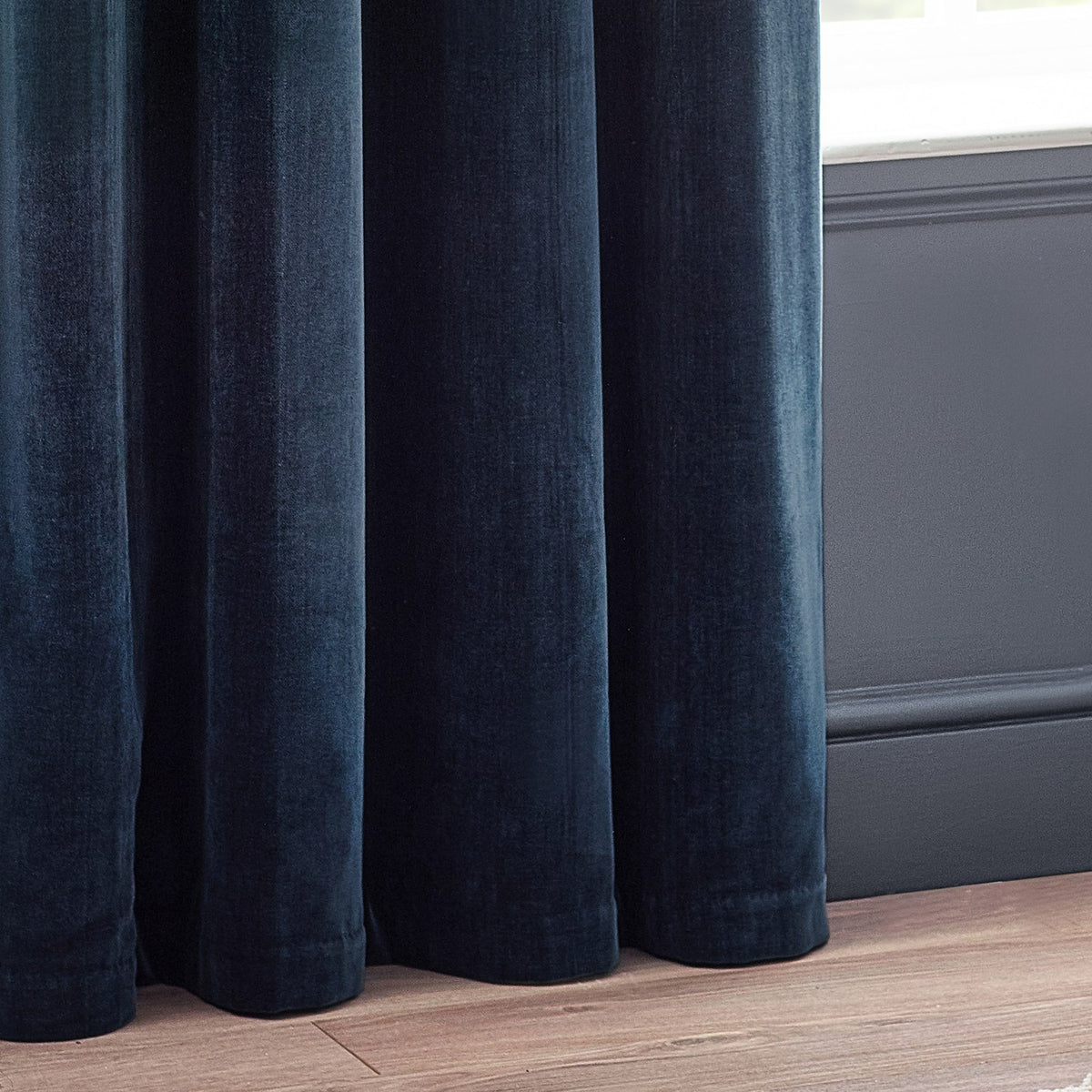 Navy heavy chenille velvet eyelet curtains with soft folds pooling on a wooden floor beside a dark panelled wall and window.