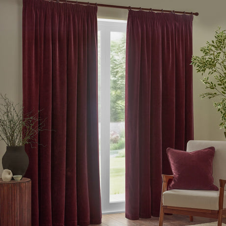 Heavy chenille velvet pencil pleat curtains in beetroot red, hung from a pole beside a window in a living room, with a matching cushion on a nearby armchair.