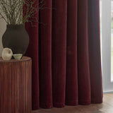 Heavy chenille velvet pencil pleat curtains in deep beetroot red, shown draped floor-length beside a window in a living room with a wooden side table.