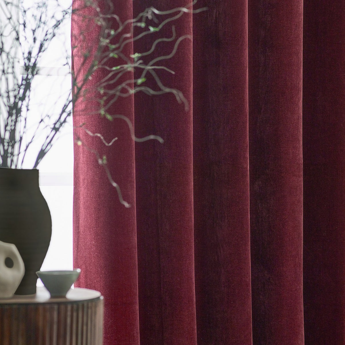 Heavy chenille velvet pencil pleat curtains in deep beetroot red, shown in folds by a window with a dark vase and branches on a side table.