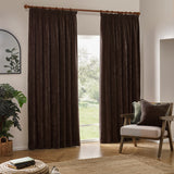 Brown heavy chenille velvet pencil pleat curtains hanging from a wooden pole in a living room, shown beside a patio door with an armchair and green plant.