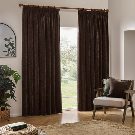 Brown heavy chenille velvet pencil pleat curtains hanging from a wooden pole in a living room, shown beside a patio door with an armchair and green plant.