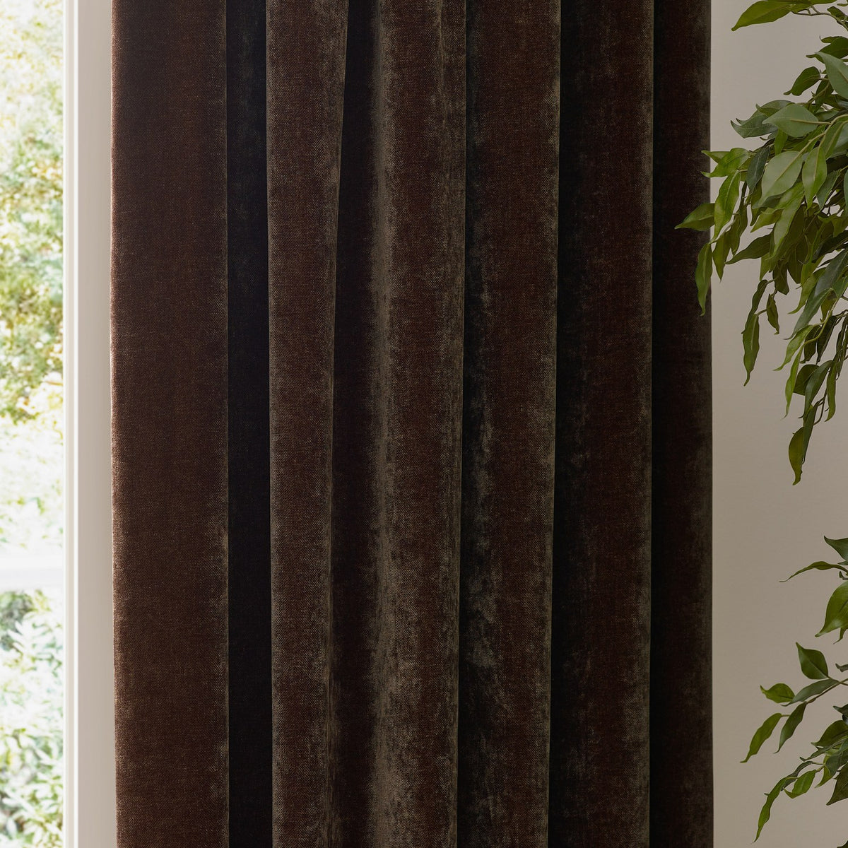 Heavy Chenille Plain Velvet Pencil Pleat Curtains in brown, showing deep folds of plush textured fabric hanging beside a window with greenery.