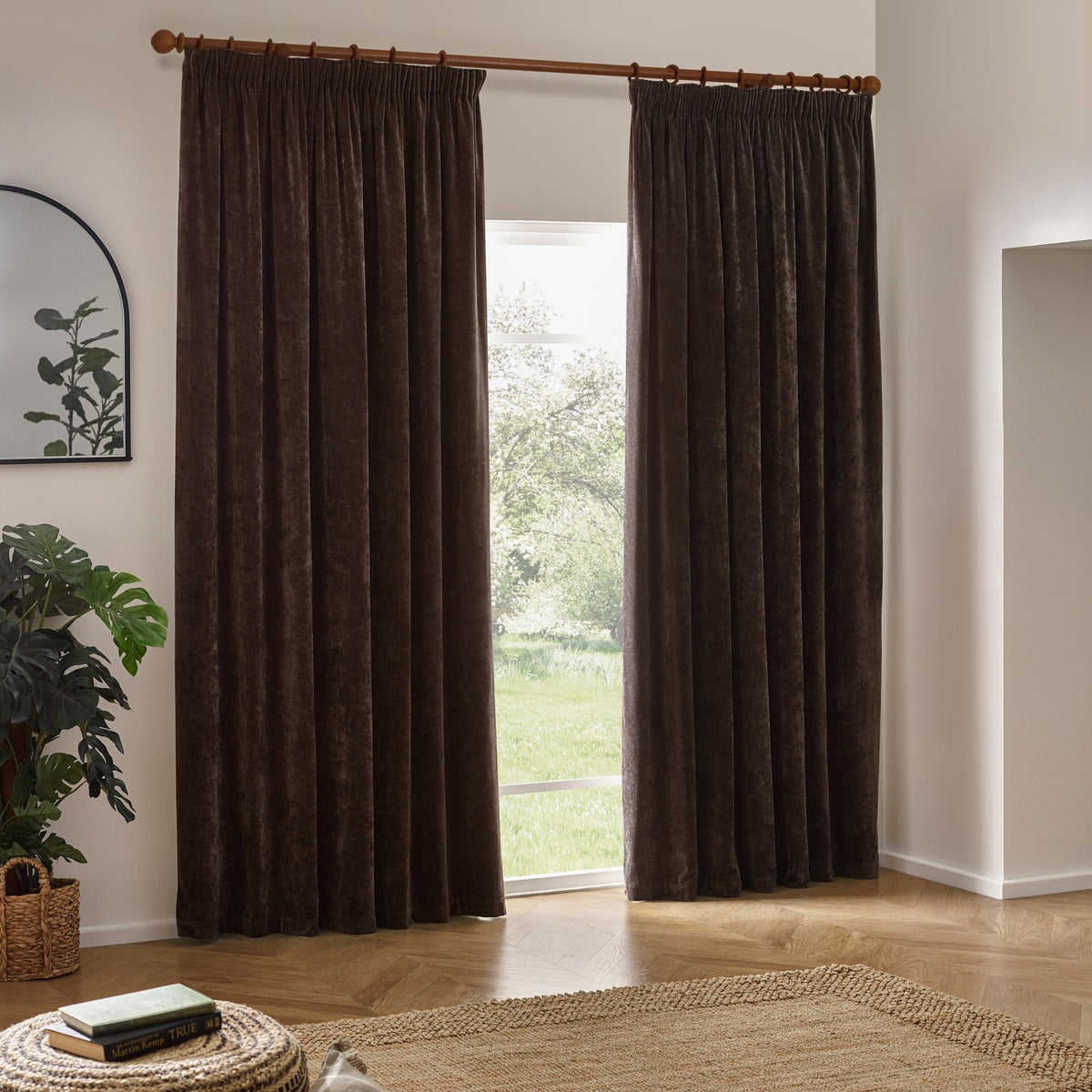 Heavy Chenille Plain Velvet Pencil Pleat Curtains in brown, hung on a wooden pole beside a bright window in a living room with wood flooring.