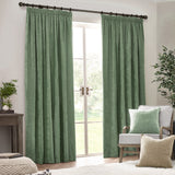 Eucalyptus green pencil pleat curtains in heavy chenille velvet, shown hung on a dark pole beside a window in a living room with an armchair and cushions.