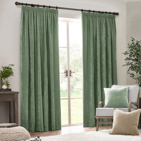 Eucalyptus green pencil pleat curtains in heavy chenille velvet, shown hung on a dark pole beside a window in a living room with an armchair and cushions.