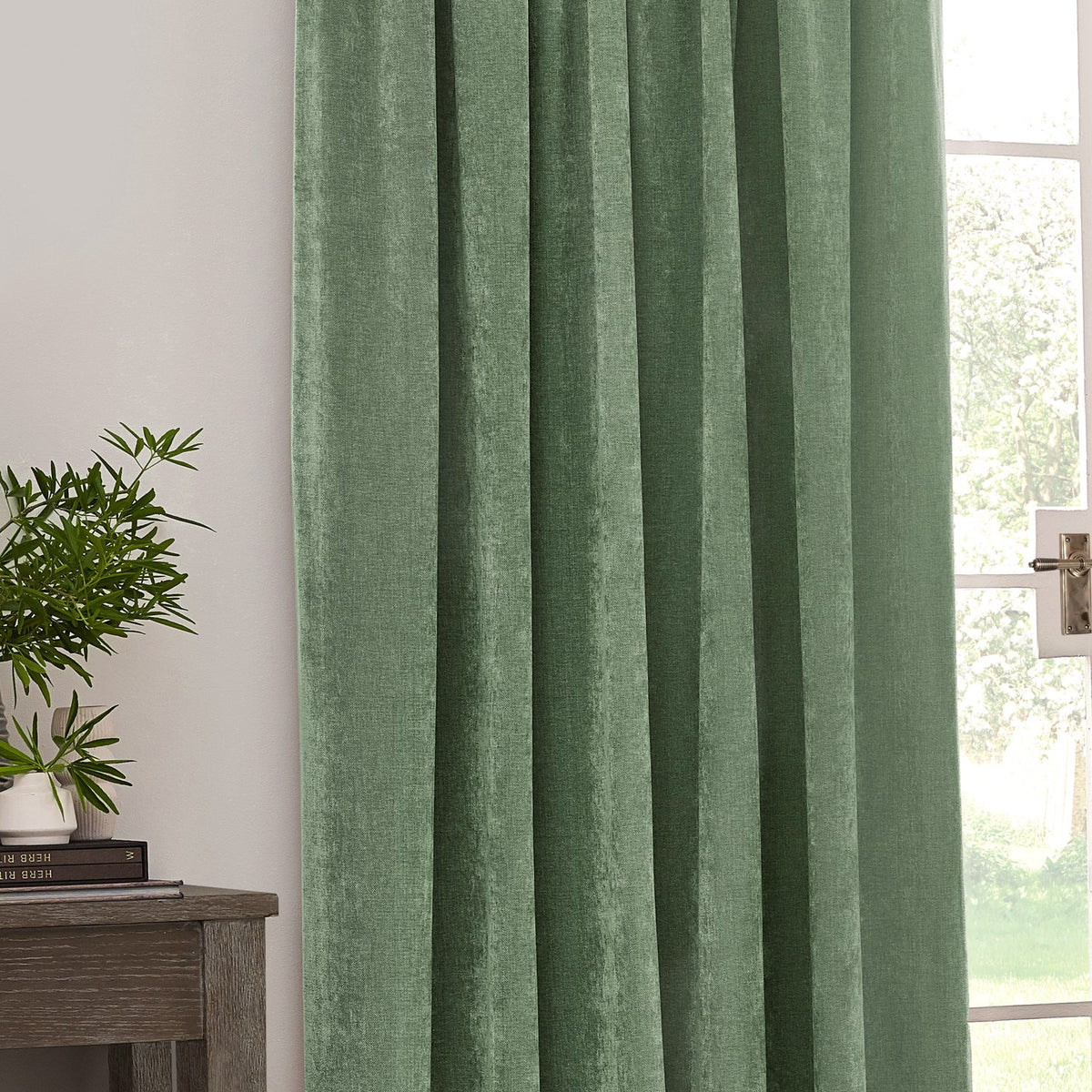 Heavy Chenille Plain Velvet Pencil Pleat Curtains in eucalyptus green, shown hanging in pencil-pleat folds beside a window in a living room setting.