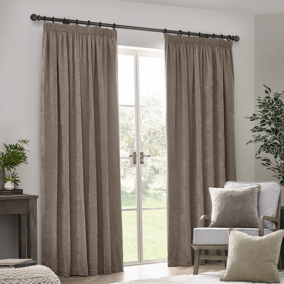 Pencil pleat curtains in greige chenille velvet hung either side of a French door on a black pole, styled in a living room with an armchair and cushions.
