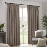 Pencil pleat curtains in greige chenille velvet hung either side of a French door on a black pole, styled in a living room with an armchair and cushions.