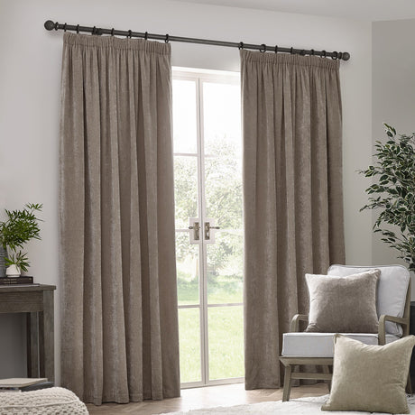 Pencil pleat curtains in greige chenille velvet hung either side of a French door on a black pole, styled in a living room with an armchair and cushions.