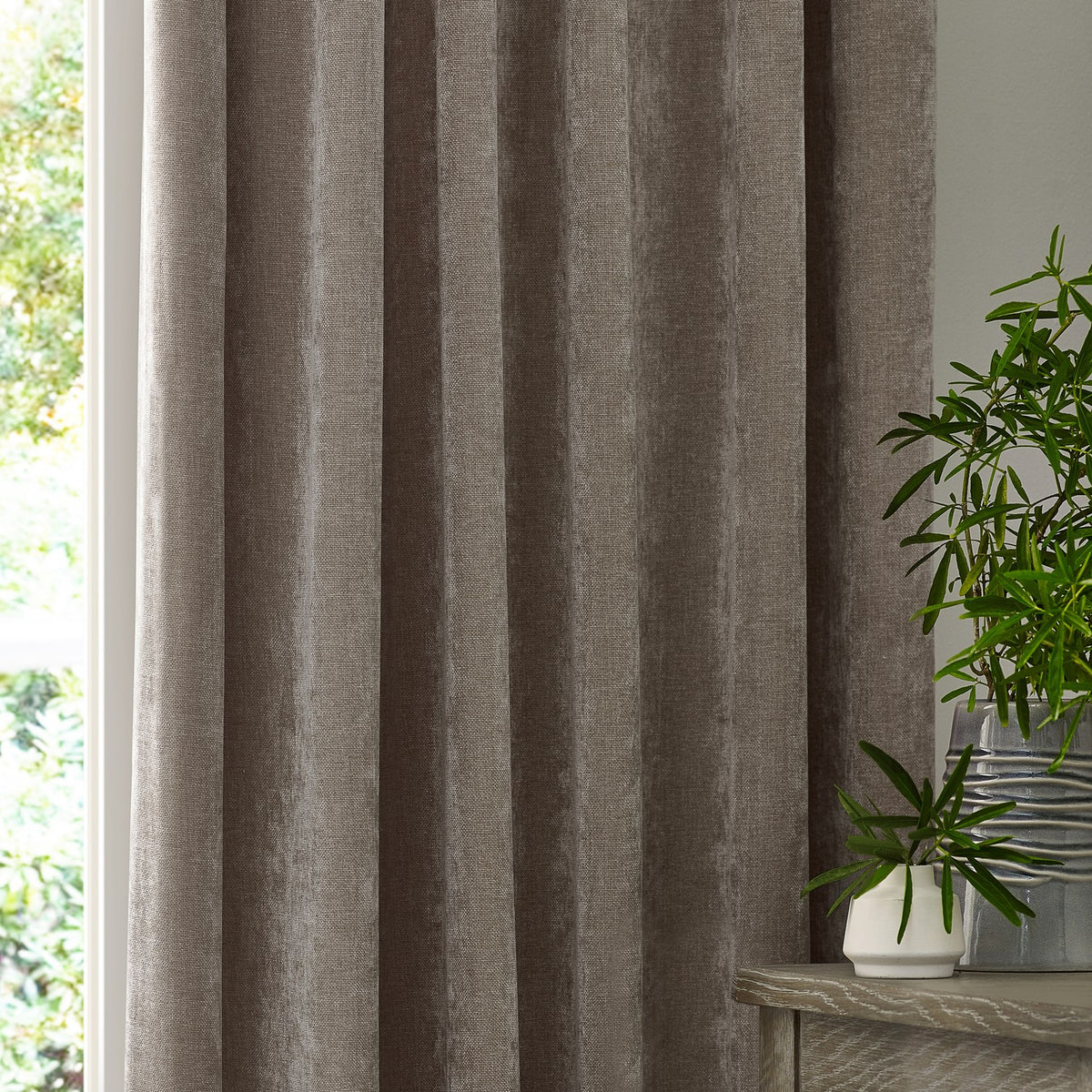 Heavy Chenille Plain Velvet Pencil Pleat Curtains in greige, shown draped in soft folds beside a window, with plants and a wooden table nearby.