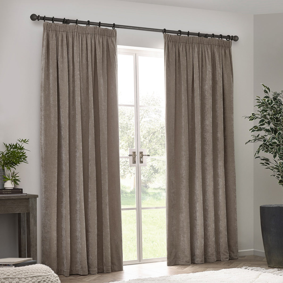 Pair of pencil pleat curtains in greige chenille velvet, hanging from a dark metal pole beside a glass door in a living room setting.