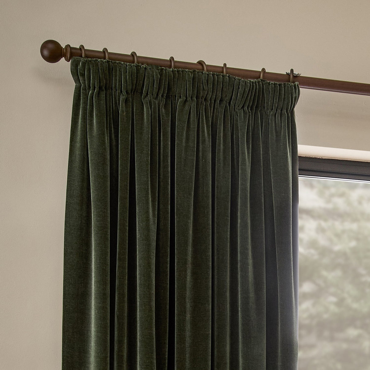 Heavy chenille velvet pencil pleat curtains in lichen green hanging from a wooden pole with rings, shown beside a window in a living room.