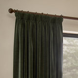 Heavy chenille velvet pencil pleat curtains in lichen green hanging from a wooden pole with rings, shown beside a window in a living room.