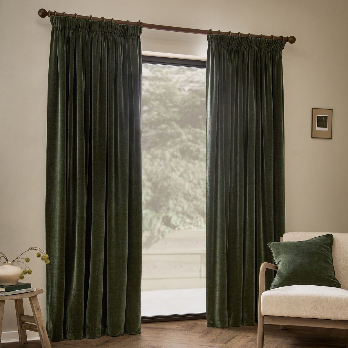 Lichen green heavy chenille velvet pencil pleat curtains hanging fully lined at a window, shown in a living room with a beige armchair, cushion, and wooden stool.