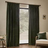 Lichen green heavy chenille velvet pencil pleat curtains hanging fully lined at a window, shown in a living room with a beige armchair, cushion, and wooden stool.